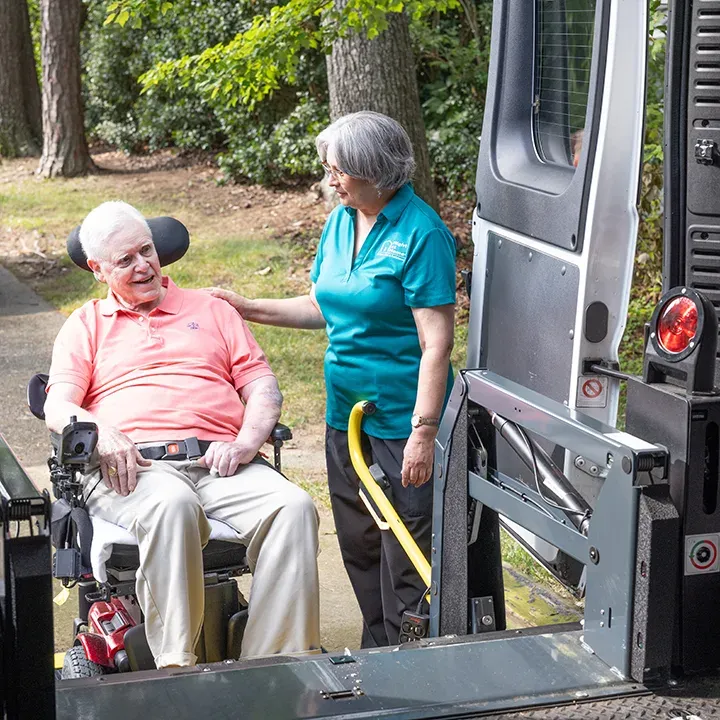 An elderly patient in a wheelchair boards a medical van with a special lift, assisted by a care provider.