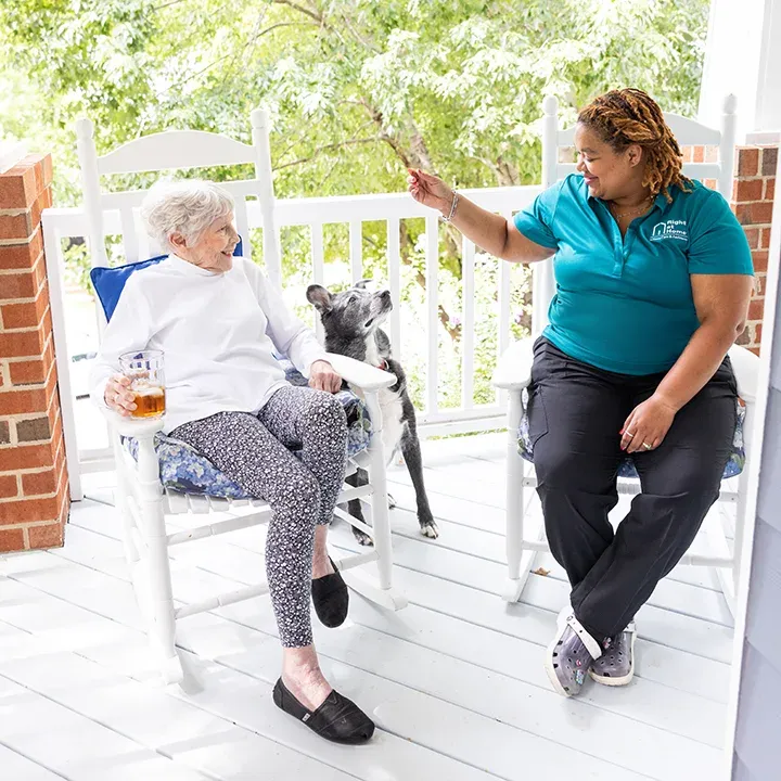A healthcare professional in blue medical scrubs spends time with an elderly patient and a dog at the front door of a home.