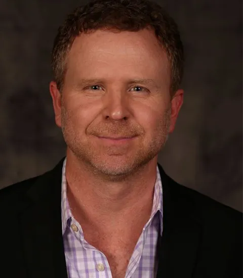 A portrait photo of a man wearing a purple checked shirt and a black jacket.