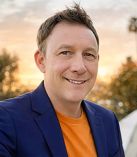 A portrait photo of a man wearing an orange shirt and a blue jacket, with a sunset visible in the background.